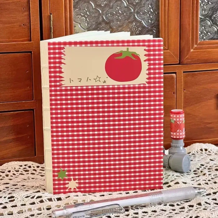 Un cuaderno de cuadros rojos con un tomate, de pie sobre un tapete de ganchillo, con un sello y un portaminas